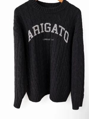 Svart stickad tröja Axel Arigato - Svart kabelstickad tröja från Axel Arigato med stor vit text 'ARIGATO' och mindre text 'LONDON '14' på bröstet. Tröjan har rund halsringning och ribbade muddar. Perfekt för dig som gillar streetwear och snygga detaljer. Storlek S men passar även M