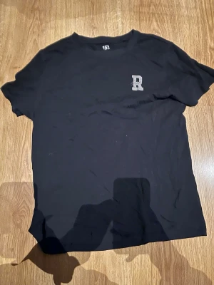 Svart t-shirt med R-tryck 157 - Svart t-shirt från 157 med ett litet grått 'R' tryckt på vänster bröst. Klassisk rund halsringning och korta ärmar. Perfekt basic-plagg för en clean och enkel stil.