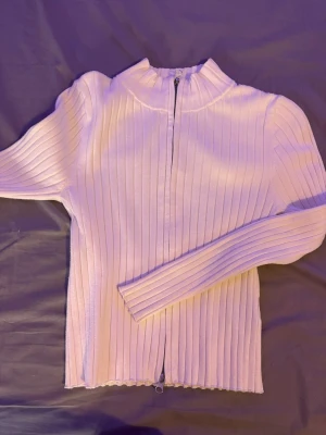 Vit ribbad ziptröja från H&M - Säljer en vit ribbad tröja från H&M med dragkedja framtill. Tröjan har lång ärm, hög krage och är tillverkad i ett stretchigt material som sitter snyggt. Perfekt till jeans eller kjol för en clean look.