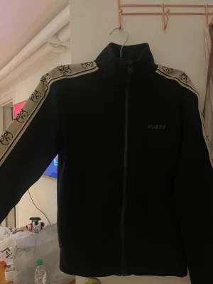 Svart ziptröja från Guess - Svart ziptröja från Guess med hög krage och dragkedja framtill. Beigea ränder med Guess-logga längs ärmarna ger en sportig vibe. Perfekt för dig som gillar streetwear och vill ha något bekvämt men ändå snyggt. Ej använd så mycket 