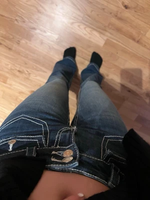 Blå bootcut jeans med kontrastsömmar - Säljer ett par blå bootcut jeans med tydliga vita kontrastsömmar och dubbla knappar fram. Jeansen har fickor både fram och bak samt en lätt tvättad look. Perfekta för dig som gillar en avslappnad men ändå trendig stil. Använt 1-2 gånger. Ny pris 500