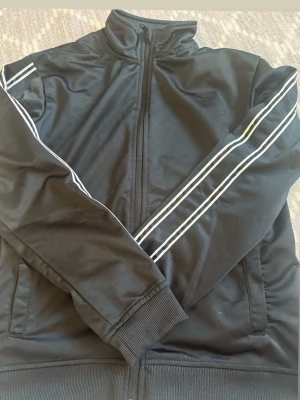 Svart track jacket med vita ränder - Snygg svart track jacket med vita dubbla ränder längs ärmarna och hög krage. Dragkedja framtill och ribbade muddar vid ärmslut och nederkant. Perfekt för en sportig och avslappnad stil.