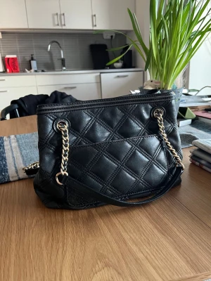 Svart quiltad handväska med kedjerem - Snygg svart handväska i quiltat mönster med guldfärgade kedjeremmar och läderdetaljer. Massor med fickor både inne och utanpå väskan. Väskan har en klassisk rektangulär form och stilrena sömmar som ger en lyxig känsla. Perfekt för dig som vill ha en trendig accessoar med plats för det viktigaste. Axelremmarna går att röra