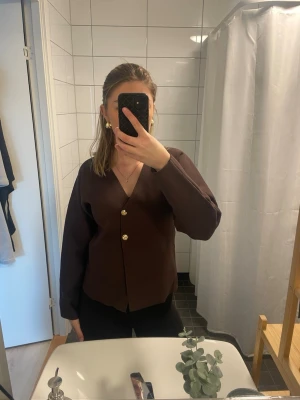 Brun/ vinröd  kofta med guldknappar från Zara - Snygg brun kofta från Zara med djup V-ringning och två dekorativa guldknappar framtill. Koftan har lång ärm och är tillverkad i ett mjukt, ribbat material som ger en skön passform. Perfekt att styla med jeans eller kjol för en trendig look.