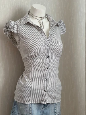 Randig blus med puffärmar - Säljer en vit blus med tunna svarta ränder och puffärmar. Blusen har knappar framtill, figurnära passform och klassisk krage. Materialet ser ut att vara bomull eller bomullsblandning. Perfekt till jeans eller kjol för en trendig look.