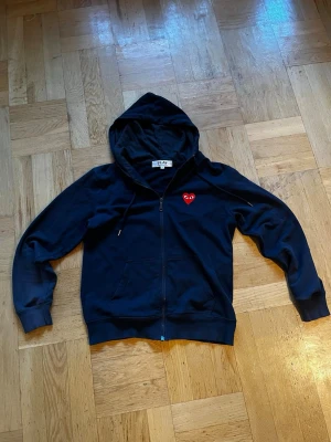 Mörkblå Comme des Garçons Play (Zip Up) - Mörkblå hoodie med dragkedja från Comme des Garçons Play. Klassisk design med huva, fickor framtill och det ikoniska röda hjärtat med ögon på bröstet. En lätt hoodie som passar för varmare väder. Storlek S men passar som XS. Säljer pga att den inte längre passar mig.