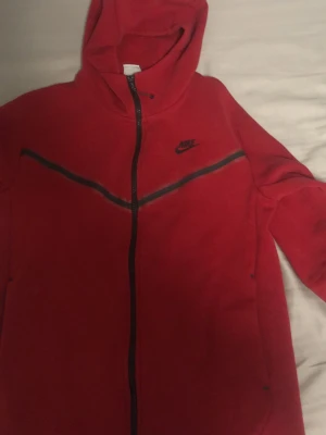 Röd Nike hoodie med dragkedja - Säljer en röd hoodie från Nike med hel dragkedja och svart Nike-logga på bröstet. Hoodien har en svart detalj i form av ett V över bröstet och huva. Tillverkad i mjukt material, perfekt för chill dagar eller träning.