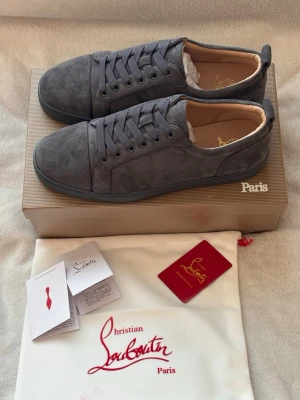 Gråa sneakers från Christian Louboutin - Stilrena gråa sneakers från Christian Louboutin i mocka med klassisk låg profil och rund tå. Skorna har matchande grå skosnören och diskreta sömmar. Insidan är fodrad i beige läder och sulan är svart. Perfekta för dig som gillar exklusiva och minimalistiska sneakers.