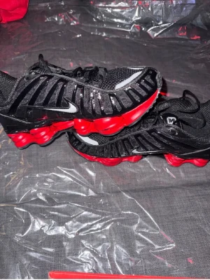 Nike Shox svarta och röda sneakers - Säljer ett par Nike Shox sneakers i svart mesh och syntet med röda fjädrande sulor. Skorna har en sportig vibe med silverfärgade swoosh-loggor på sidorna och coola detaljer längs ovandelen. Perfekta för dig som vill sticka ut med en unik och futuristisk look.