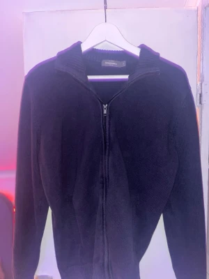 Svart ziptröja från Dressmann - En svart stickad ziptröja från Dressmann med ribbad krage och långa ärmar. Tröjan har hel dragkedja framtill och är tillverkad i ett mjukt material som passar perfekt för lager-på-lager. Enkel och stilren design.