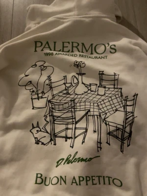 Vit hoodie med tryck  - Vit hoodie med huva och känguruficka från Palermo's. På ryggen finns ett stort tryck med en tecknad bild av ett dukat bord, gröna texter och signatur. Framtill står det 'PALERMO'S 1998 Awarded Restaurant' i grönt. Perfekt för dig som gillar unika prints.