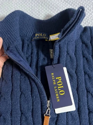 Blå kabelstickad tröja Polo Ralph Lauren - Mörkblå kabelstickad tröja från Polo Ralph Lauren med halv dragkedja och hög krage. Tröjan har långa ärmar, ribbade muddar och en liten röd broderad logga på bröstet. Klassisk och stilren design i mjukt material.OBS! storleken är L men passar som S!