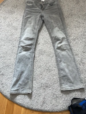 Grå bootcut jeans - Säljer ett par ljusgrå bootcut jeans med snygg ficka bak som har knapp och broderad detalj. Jeansen har klassisk femficksmodell och är i stretchigt denimtyg för skön passform. Säljer pga att de är försmå. Storlek 158. 