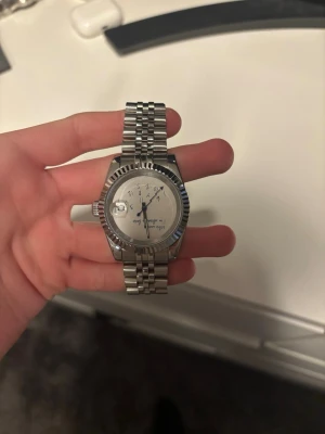 Silverfärgad klocka med länkarmband - Stilren klocka i silver med klassiskt länkarmband och räfflad bezel. Urtavlan är ljus med tydliga markeringar och datumfönster. Perfekt accessoar för dig som gillar en tidlös look.