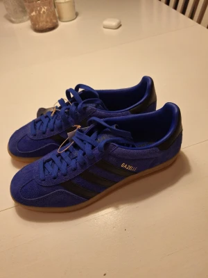 Blå Adidas Gazelle sneakers nya med prislapp. - Snygga blå Adidas Gazelle sneakers i mocka med klassiska svarta ränder på sidorna och gummisula.  Nya med prislapp. 