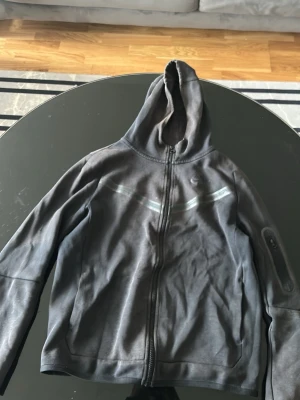 Mörkgrå Nike zip hoodie - Snygg mörkgrå hoodie från Nike med dragkedja framtill och huva. Tröjan har ett diskret Nike-märke på bröstet och reflekterande detaljer över bröstet. Tillverkad i mjukt material som passar perfekt till en avslappnad streetwear-look.