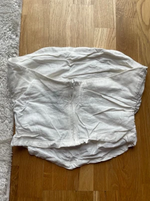 Vit axelbandslös tubtopp i linne - Säljer en vit axelbandslös tubtopp i linne. Toppen har en bandeau-form och dragkedja bak.