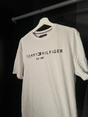 Vit T-shirt från Tommy Hilfiger - Snygg vit T-shirt från Tommy Hilfiger med klassisk logga och texten 'EST. 1985' på bröstet. T-shirten har rund halsringning med en diskret randig detalj och är kortärmad. Tillverkad i mjuk bomull för en skön känsla.