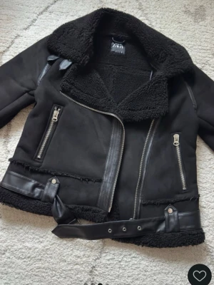 Svart teddyjacka från Zara - Snygg svart teddyjacka från Zara med stora kragar, dragkedjor och bälte i midjan. Jackan har mjukt teddyfoder, detaljer i fuskskinn och två framfickor med dragkedja. Perfekt för kalla dagar och ger en cool look.