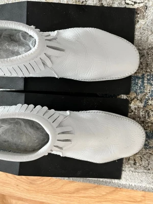 Vita mockasiner minnetonka - Unika vita boots i mjukt läder med dekorativa fransar runt skaftet och handsydda detaljer. Skorna har en klassisk mockasinform och låg klack, vilket ger en cool och avslappnad look. Perfekta för dig som vill sticka ut med din stil.