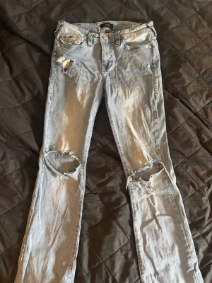 Ljusblå ripped jeans True Religion - Säljer ett par ljusblå jeans från True Religion med stora slitningar vid knäna och mindre hål upptill. Klassisk femficksmodell med raka ben och broderad bakficka. Jeansen är i denim och har en avslappnad, trendig look.