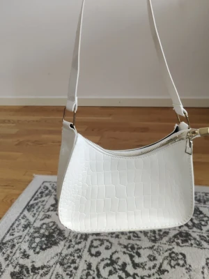 Vit axelväska - Snygg vit axelväska yta och justerbar axelrem. Väskan har en böjd form och stängs med dragkedja upptill. Perfekt för dig som gillar stilrena accessoarer med lite edge.