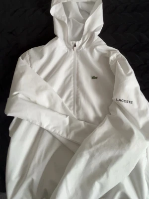 Vit hoodie från Lacoste med dragkedja - Snygg vit hoodie från Lacoste med dragkedja framtill och klassisk huva. Diskret Lacoste-logga på bröstet och texten 'LACOSTE' på ärmen. Tillverkad i ett lätt och mjukt material som känns skönt mot huden. Perfekt för en clean och sportig look.