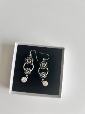 Silvriga örhängen med rosor och hängande detaljer - Säljer ett par coola silvriga örhängen med dubbla rosor och cirklar. De har en hängande rund detalj längst ner som ger extra touch. Örhängena är i metall och har krokar för enkel användning. Perfekt för dig som gillar unika accessoarer med blommönster.