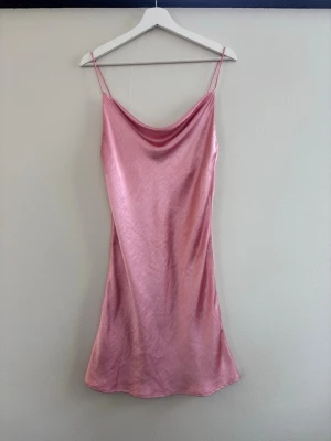 Rosa satin slipklänning - Säljer en rosa slipklänning i glansigt satinmaterial med tunna axelband och mjukt draperad urringning. Klänningen har en kort längd och en enkel, elegant siluett som ger en trendig och feminin look.