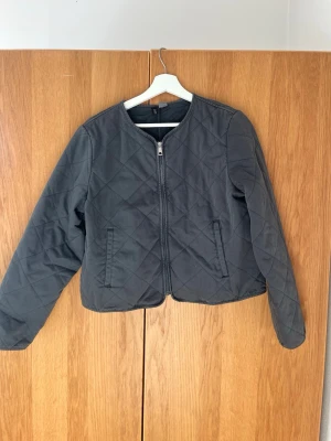 Svart quiltad pufferjacka - Snygg svart/grå pufferjacka med quiltat mönster och dragkedja framtill. Jackan har två fickor på sidorna och en rak, kortare passform. 