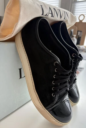 Svarta Lanvin sneakers i mocka och lack - Snygga svarta sneakers från Lanvin med ovandel i mocka och tåparti i blankt lack. Skorna har vita sulor, svarta skosnören och metallfärgade öljetter. Perfekta för dig som gillar stilrena och exklusiva sneakers med en twist.