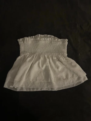 Vit ärmlös bandeau topp med volang - Supersöt vit bandeau topp med smockad överdel och volangkant upptill. Toppen är ärmlös och har ett lätt prickigt mönster i tyget samt en luftig, flowy nederdel. Perfekt för varma dagar och somriga outfits.