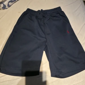 Mörkblå shorts från Polo Ralph Lauren - Säljer ett par mörkblå shorts från Polo Ralph Lauren med elastisk midja och det klassiska röda logobroderiet på benet. Sköna och stilrena, perfekta för sommaren. Passar till allt och har en avslappnad passform.
