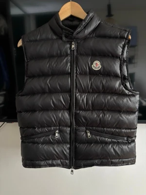 Svart dunväst från Moncler - Stilren svart dunväst från Moncler med hög krage och två dragkedjeförsedda fickor framtill. Västen har ett quiltat mönster och Moncler-logga på bröstet. Perfekt att slänga över en hoodie eller tröja för extra värme och stil. Storlek 1 vilket motsvarar S