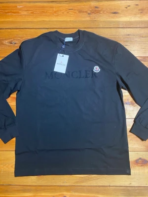 Svart långärmad tröja från Moncler - Snygg svart långärmad tröja från Moncler med diskret logotyp på bröstet och broderad Moncler-patch på vänster sida. Tröjan har rund halsringning och ribbade muddar vid ärmslut. Perfekt för en clean och stilren look.