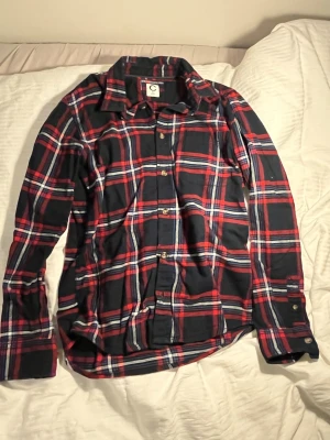 Rutig skjorta i rött och blått från C - Snygg långärmad skjorta från C i klassiskt röd, blå och vit rutigt mönster. Skjortan har knappar framtill och vid ärmsluten samt en mjuk bomullskänsla. Perfekt för dig som gillar en avslappnad men stilren look.