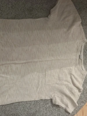 Ljusbeige t-shirt med bröstficka - En enkel och stilren ljusbeige t-shirt med korta ärmar och en liten bröstficka. Perfekt basplagg till garderoben, gjord i mjuk bomull för skön känsla. Passar till allt och är lätt att matcha med andra plagg.