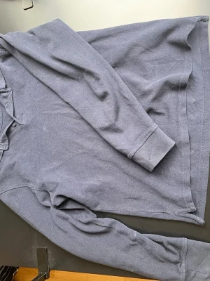 Mörkblå långärmad tröja  - Mörkblå långärmad tröja från 157 med klassisk krage och knappar vid halsen. Tröjan är i mjukt material och har ribbade muddar vid ärmslut och nederkant. Perfekt för lager-på-lager och enkel att matcha med jeans eller chinos.