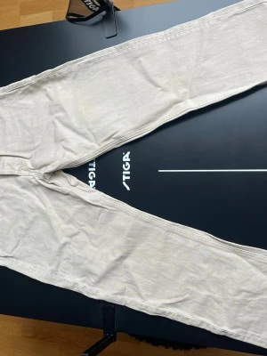 Beiga raka jeans med hög midja - Snygga beiga jeans med rak passform och hög midja. Klassisk femficksmodell och bälteshällor. Jeansen har en patch i läderimitation bak i midjan och är tillverkade i ett mjukt bomullstyg. Perfekta för en avslappnad och trendig look. Använda en gång