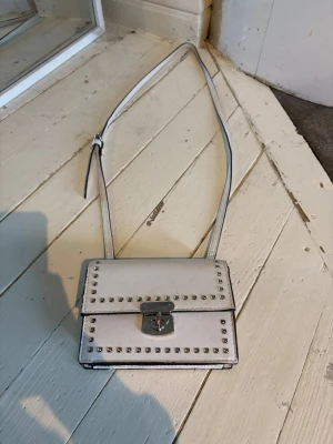 Vit axelväska med nitar - Snygg vit axelväska i boxig modell med silverfärgade nitar runt locket och ett metallspänne framtill. Justerbar axelrem och stilren design som passar till många outfits. Väskan är gjord i ett slätt konstlädermaterial.