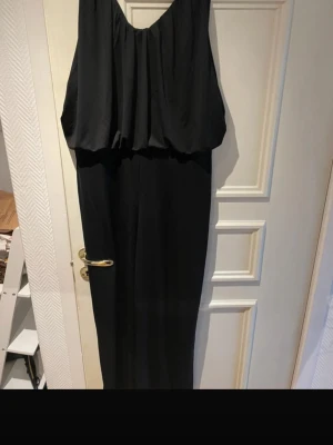 Svart långklänning i polyester - Elegant svart långklänning med lös passform och draperad överdel. Klänningen har ärmlös design och är tillverkad i mjuk polyester som faller snyggt. Perfekt för dig som gillar stilrena och tidlösa plagg.