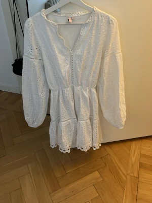 Vit kort klänning med broderi - Söt vit kortklänning i bomull med broderade detaljer och volangkant. Klänningen har långärmade ärmar, v-ringning och fina spetsdetaljer längs kanterna. Perfekt för sommarens alla tillfällen.