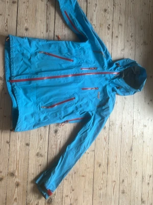 Blå regnjacka från Bergans, M - Snygg blå regnjacka från Bergans med röda dragkedjor och huva. Jackan har flera praktiska fickor med dragkedja och justerbara ärmslut. Perfekt för regniga dagar och outdoor-äventyr. Tillverkad i slitstarkt, vattentätt material.