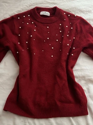 Röd stickad tröja med pärlor H&M - Snygg röd stickad tröja från H&M med dekorativa vita pärlor på framsidan. Tröjan har rund halsringning, långa ärmar och ribbade muddar vid ärmslut och nederkant. Perfekt för jul eller andra festligheter 