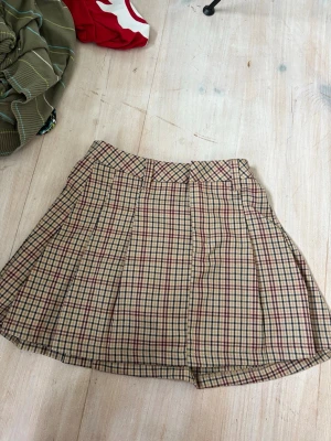 Rutig kortkjol från H&M Divided - Säljer en rutig kortkjol från H&M Divided i beige, rött och mörkblått. Kjolen har klassiskt rutmönster och är lätt utställd med bred linning. Perfekt för dig som gillar preppy stil och vill ha en kjol som sticker ut lite extra.