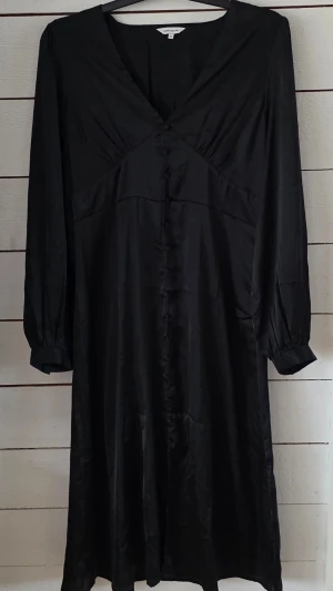 Svart långärmad klänning Bubbleroom - Stilren svart klänning från Bubbleroom med v-ringad hals, långa ärmar och knappar framtill. Klänningen har en lätt glansig finish och är gjord i ett mjukt material som ger en elegant look. Perfekt för dig som gillar klassiska och tidlösa plagg.