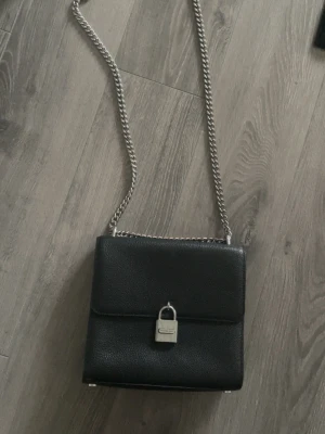 Svart axelväska med kedjerem - Snygg svart axelväska med silverfärgad kedjerem och ett coolt lås framtill. Väskan har en stilren, fyrkantig form och passar perfekt för dig som gillar minimalistisk design och trendiga detaljer.