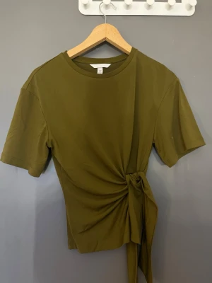 Olivgrön topp med knyt i sidan - Snygg olivgrön topp med rund halsringning och korta ärmar. Toppen har en cool knytdetalj i sidan som ger en unik twist till outfiten. Sitter snyggt på kroppen och är sååå snygg. Basic men jättefint