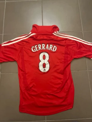 Liverpool Gerrard matchtröja Adidas - Klassisk röd Liverpool FC matchtröja från Adidas med Gerrard och nummer 8 på ryggen. Tröjan har vita detaljer, klubbmärke och Carlsberg-logga på bröstet samt vita ränder på axlarna. Perfekt för fotbollsfans och samlare. Den är i size 152 cm