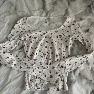 Vit blommig långärmad topp från Nelly - Supersöt vit långärmad topp från Nelly med små vinröda blommor och hjortmotiv. Toppen har en rundad halsringning med spetskant och är figurnära i modellen. Perfekt för dig som gillar romantiska och gulliga detaljer. slutsåld!